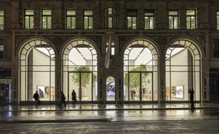 Exterior view of Apple’s London flagship store with the lights on in the evening. The store features large arched windows offering a view into the store and there are four people on the street outside