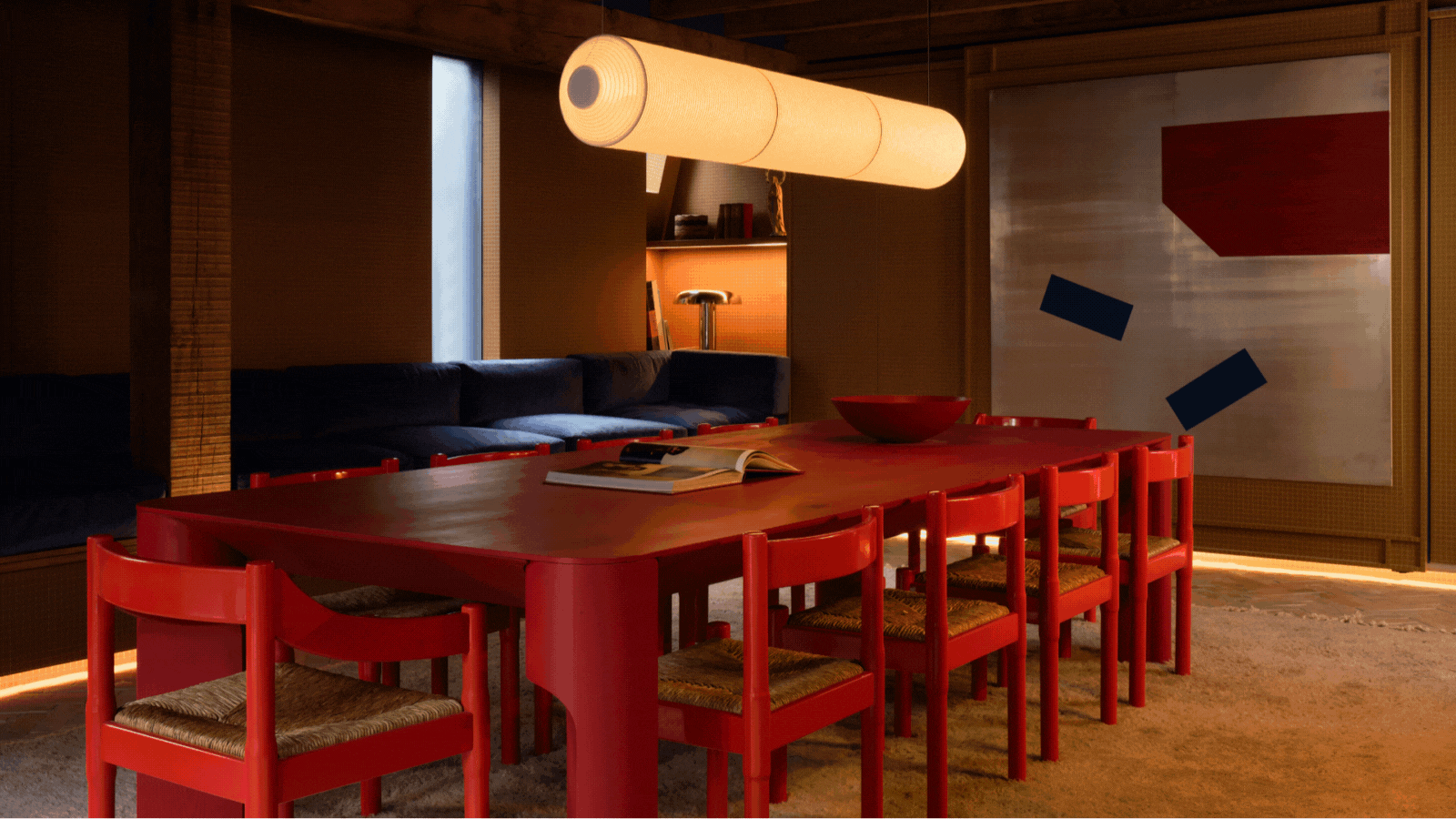 GIF of two images of a dining room that is ochre yellow with a large red dining table in the middle and a long and low navy blue bench seating to the side.