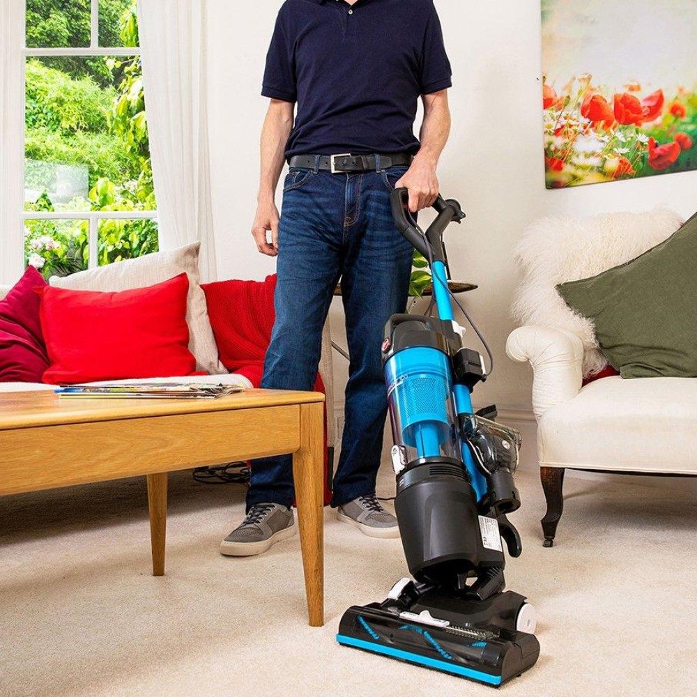 Standing man in lounge holding Hoover Upright 300 Pets with white couches with red cushions and light brown coffee table