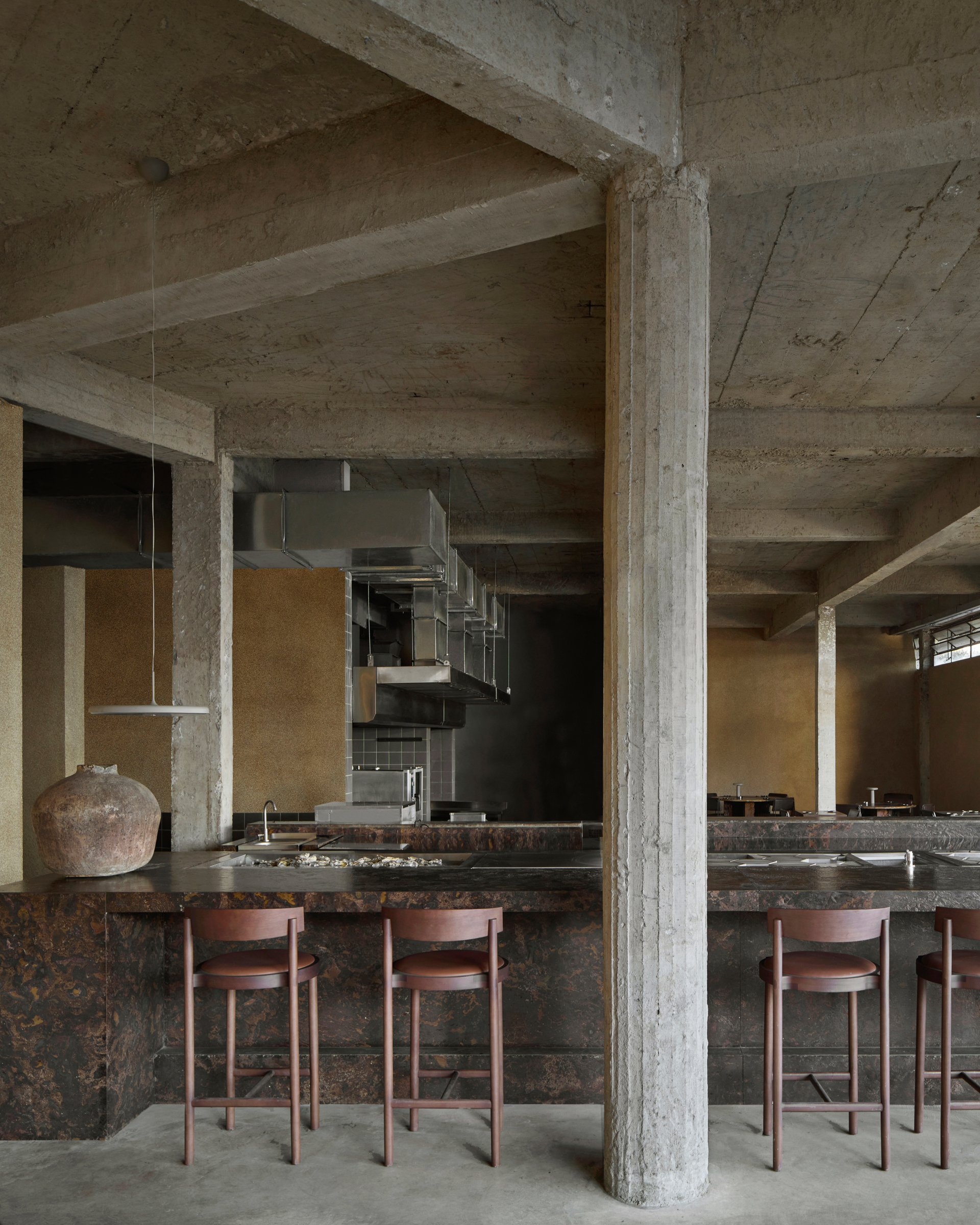 A brutalist building houses a sleek, almost utopian restaurant crafted from chocolatey slabs of stone, sheeny chrome, and exposed cement columns, with organically shaped, back-lit sconces resembling rocks.