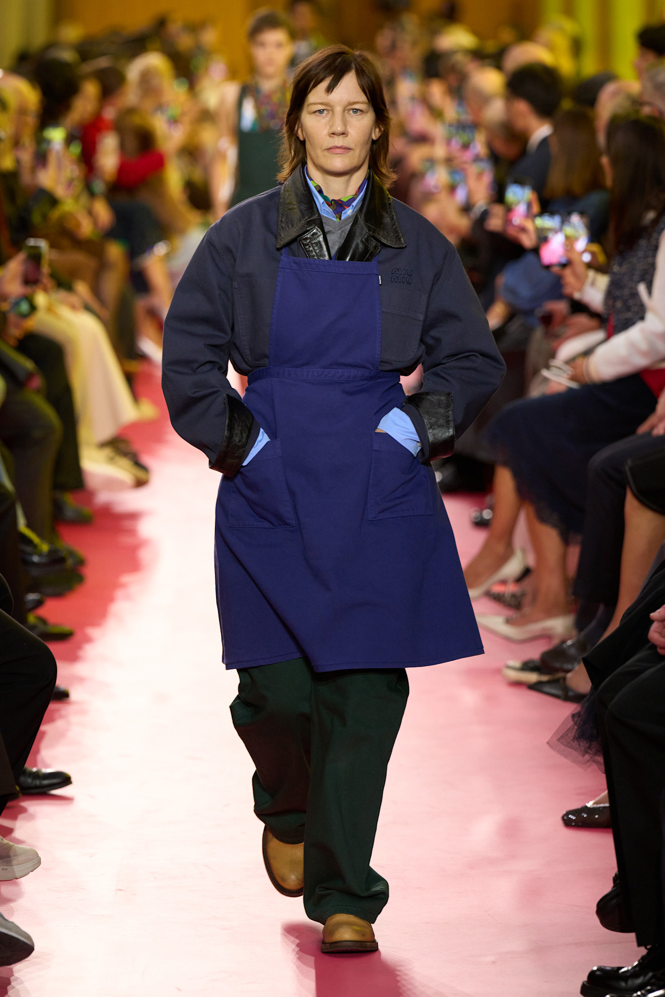 Sandra H&uuml;ller wearing a blue apron, blue barn jacket, green pants, tan boots while walking in Miu Miu's Spring 2026 runway show.