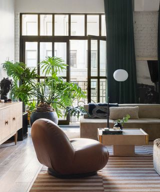 Rounded chestnut colored brown leather sofa in small living room with potted palm, looking out to balcony with black-framed doors