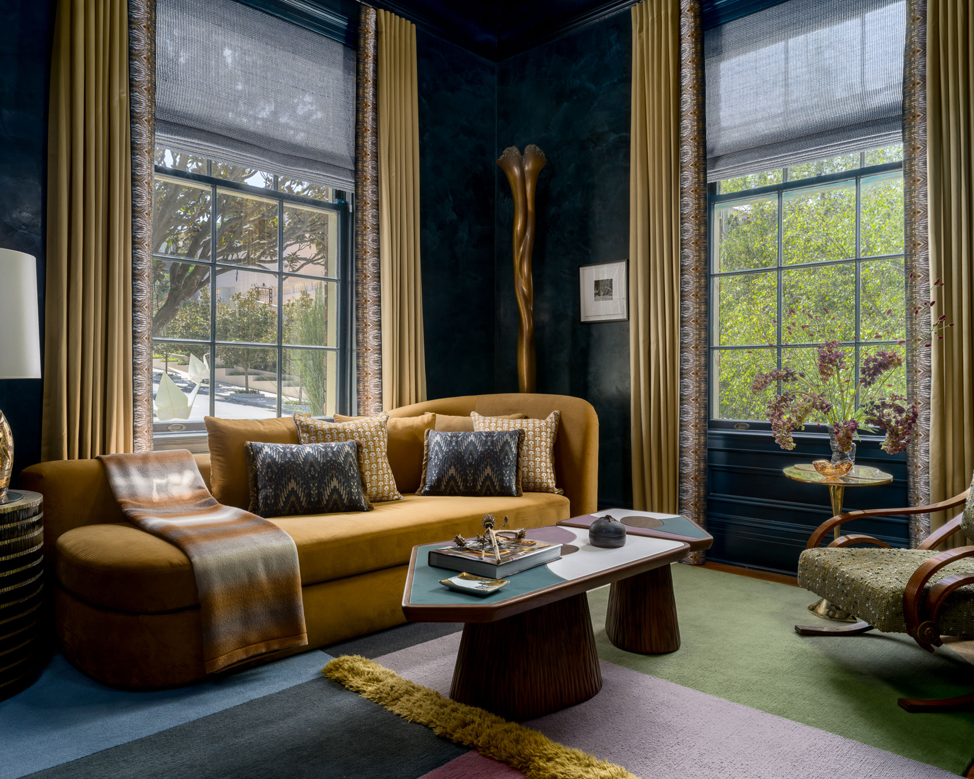 A moody, eclectic living room with navy plaster walls, a mustard velvet sofa, and geometric coffee tables. Large windows with gold drapes frame lush greenery, all set on a colorful, paneled rug.