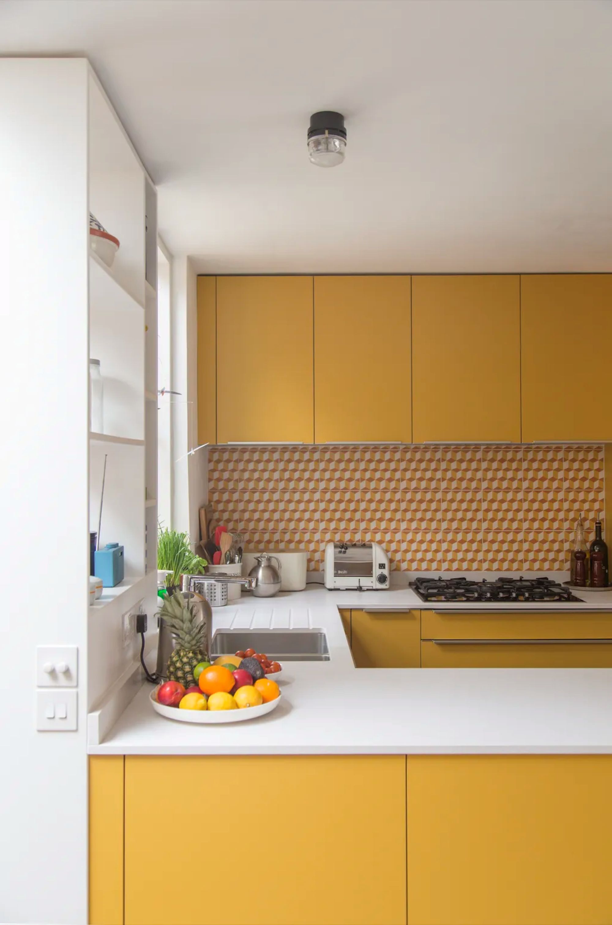 A kitchen with bold yellow cabinets, a geometric orange and white backsplash, white countertops, and fruit on top of the counter