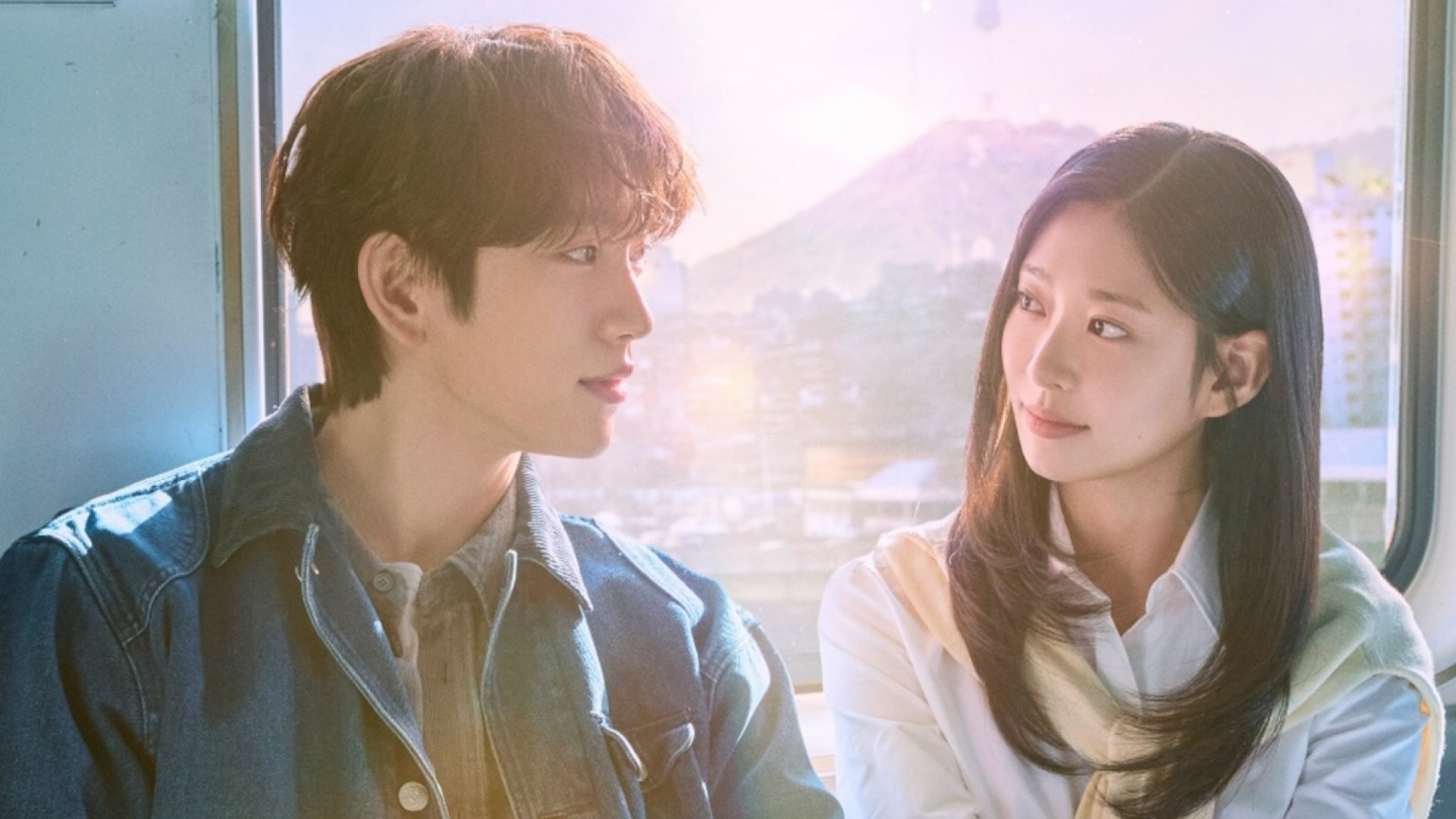 A Korean man (Park Jin-young) and a Korean woman (Kim Min-ju) look at each other as they sit in front of an overground subway car's window, with a sunny Seoul landscape behind them, in a promo image for the K-drama 'Still Shining.'