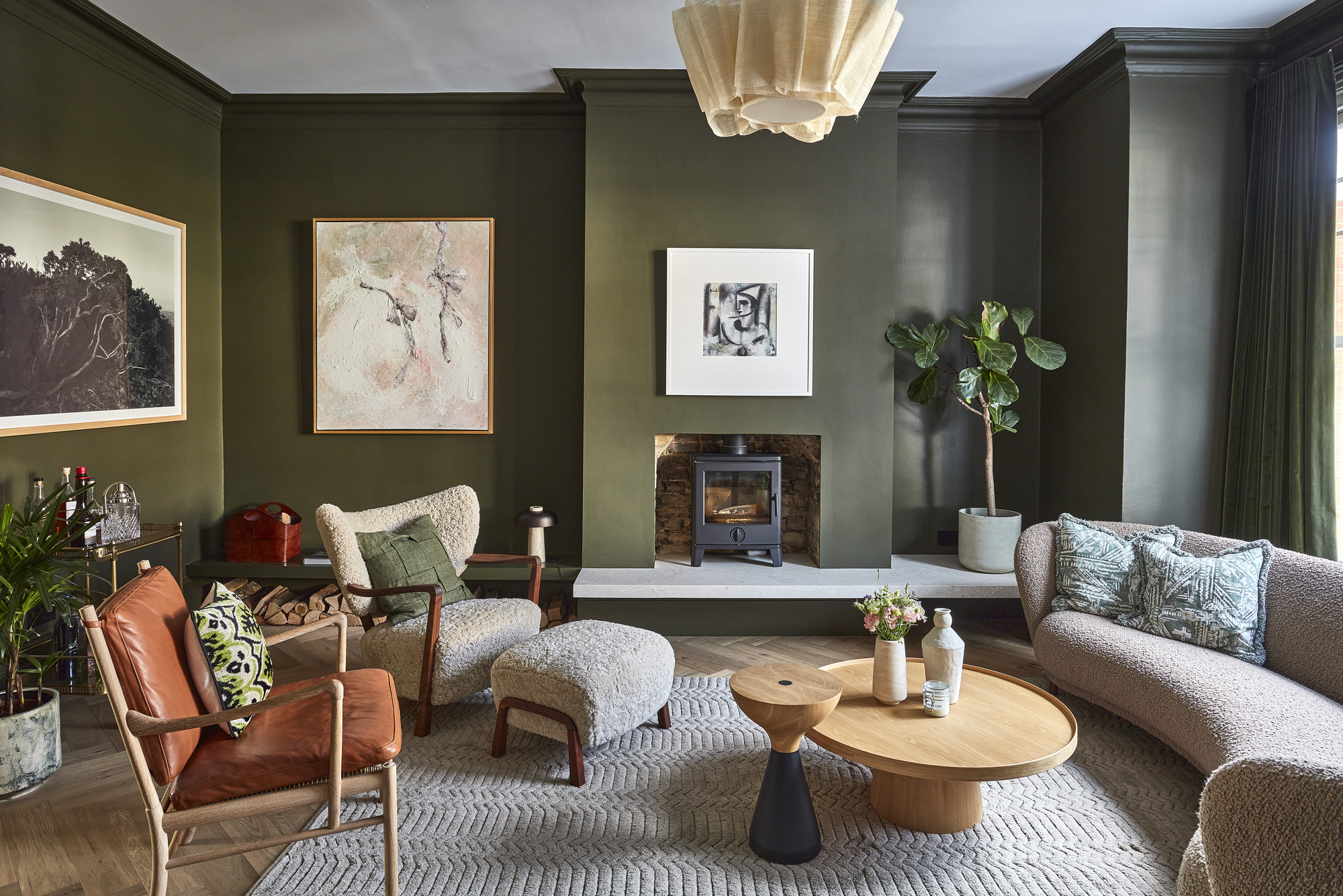 living room with dark green walls, fireplace on raised platform, artwork hung on walls, white ceiling, timber parquetry floors, a boucle armchair with matching ottoman, a tan leather and timber armchair, a round timber coffee table and side table, and a curved sofa opposite, all on a gray rug