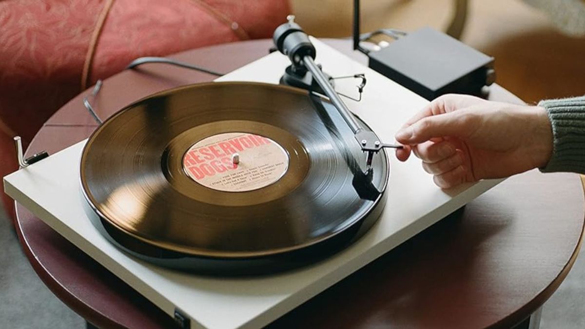 The white Pro-Ject E1 record player on a table with the vinyl of the Reservoir Dogs soundtrack on the plinth. A man&#039;s arm is positioning the needle on the first track