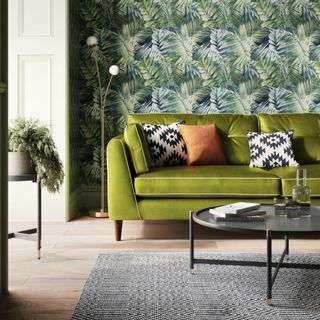 A living room with a green velvet sofa with contrasting white piping against a botanical-print wallpaper on the wall