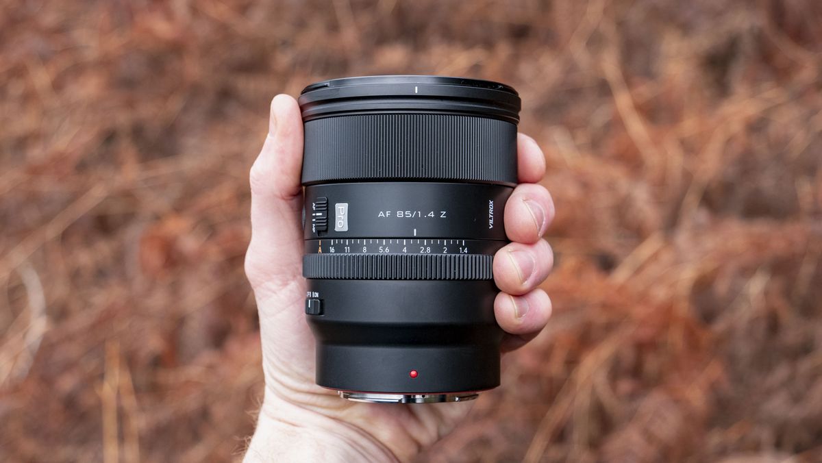 Viltrox AF 85mm F1.4 Pro lens in photographer's hand, outdoors and with bronze-colored ferns in the background