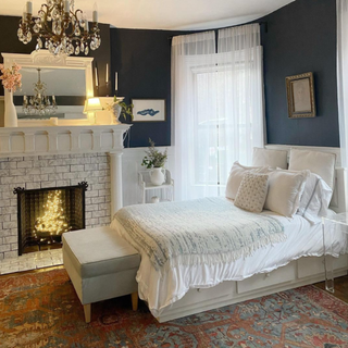 Classic bedroom scheme with dark walls, white storage bed, upholstered bench at foot end, vintage chandelier, grand tiled fireplace with mirror on mantel, and illuminated fairy lights bundle in fireplace.