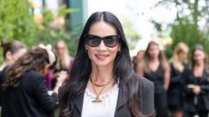 Lucy Liu wearing sunglasses, a white top, black jacket and gold chain necklaces