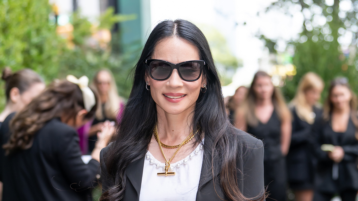 Lucy Liu wearing sunglasses, a white top, black jacket and gold chain necklaces