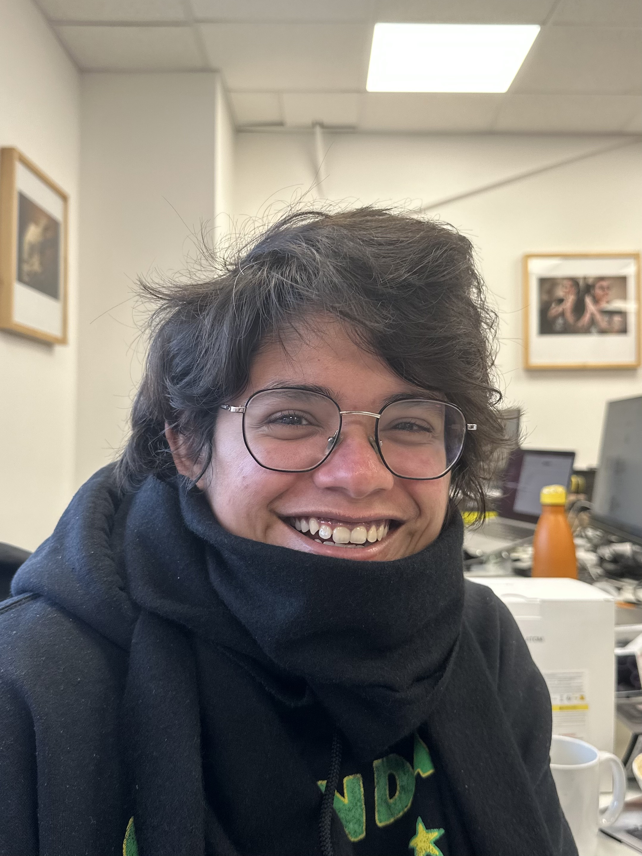 A photo of a person smiling in an office, with a black scarf around their neck and glasses.