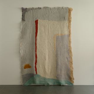 A large-scale textile creation features uneven swathes of grey, red, aquamarine, orange, lavender, and pink whilst hanging against a white background.