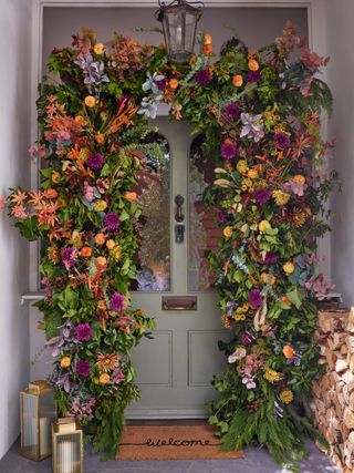 sage green front door with fall floral arch, log pile, door mat, two brass lanterns