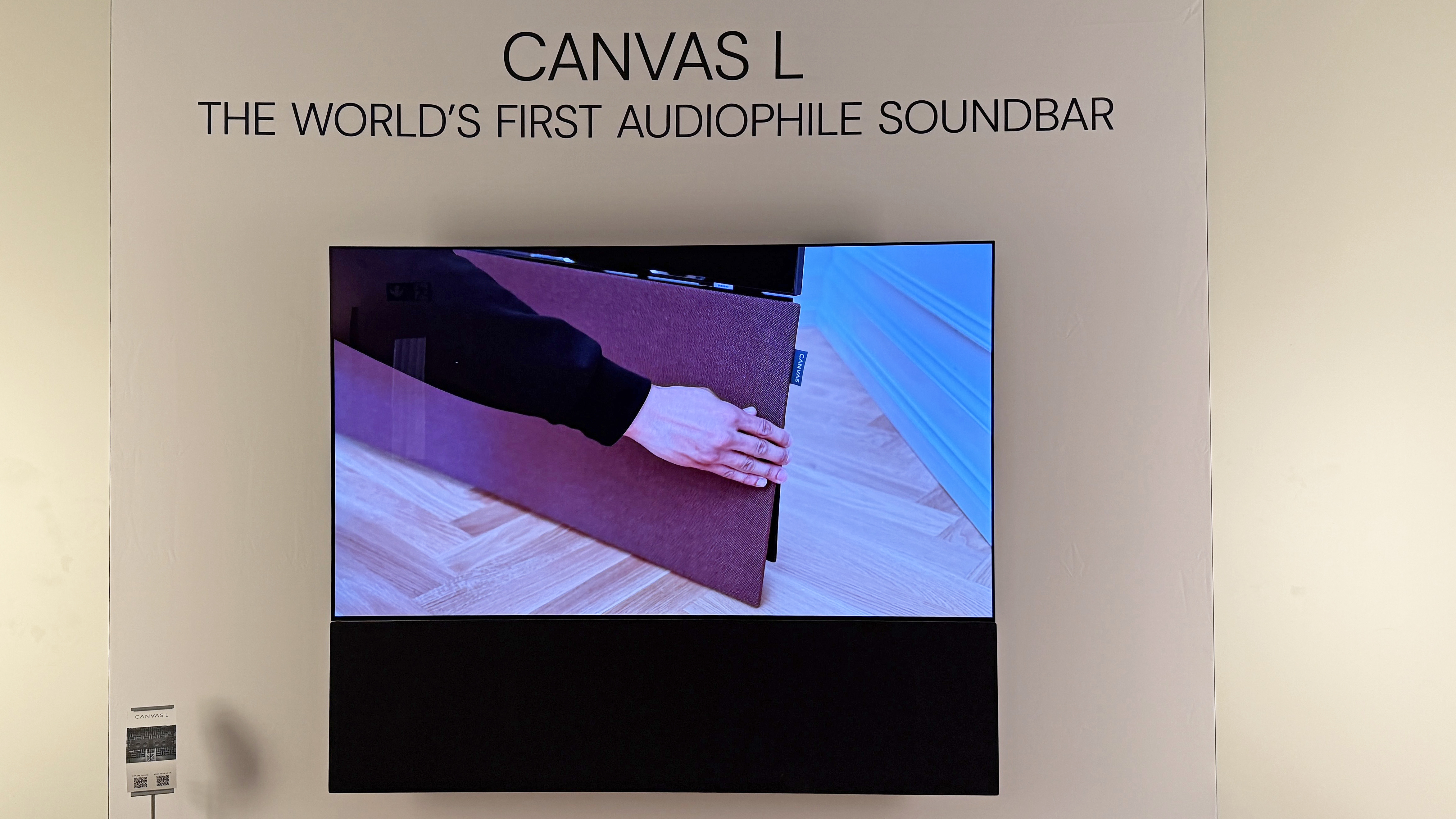 The Canvas L soundbar mounted on a wall with a TV above it. The TV is showing someone removing the grille from the Canvas soundbar