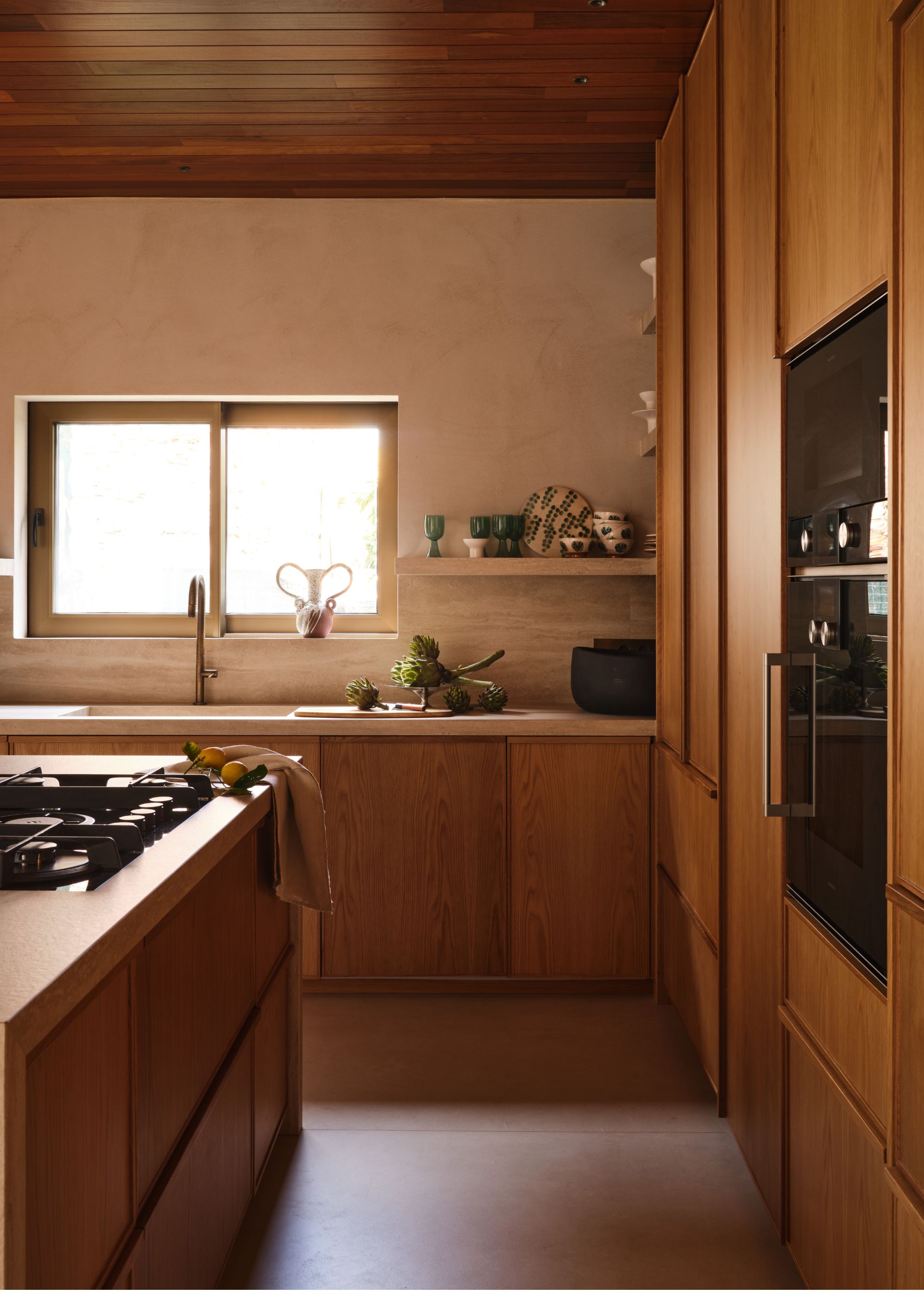A modern wood kitchen with an island hob, integrated storage and an open shelf with decorative dinnerware and glasses next to the window
