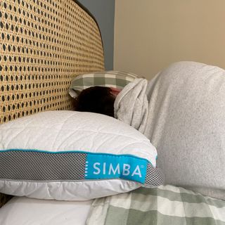 The Simba Hybrid pillow being slept on by a man with dark hair and a grey hooded swearshirt on a bed with a green and white check duvet