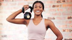 Smiling woman holding kettlebell on her shoulder, in front of a brick wall