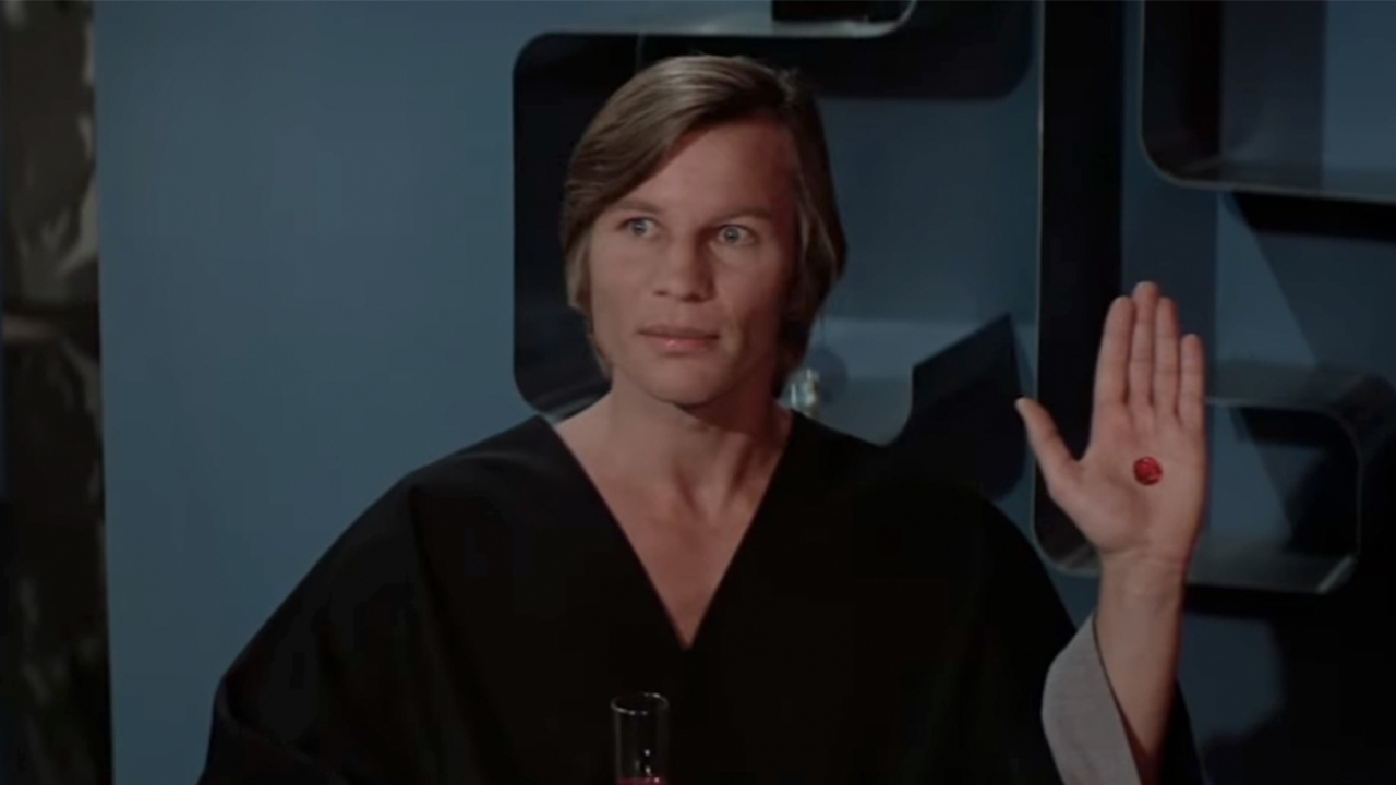 Michael York holding up his left hand with a red crystal embedded in it. 