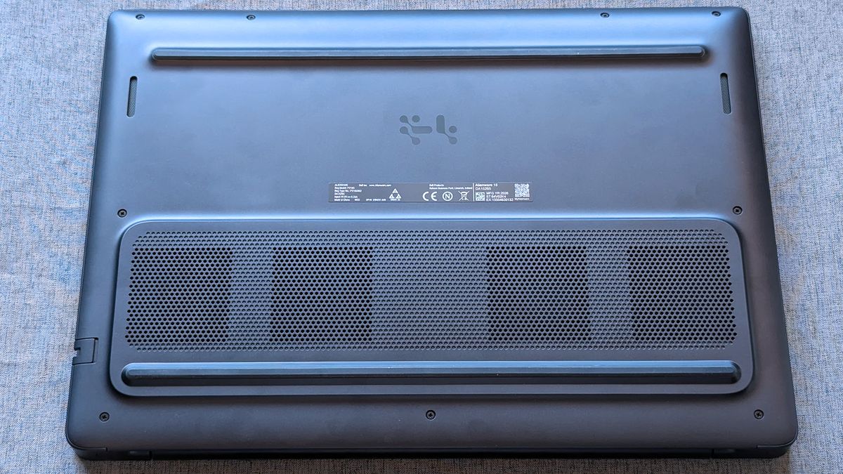 The underside of an Alienware laptop rests on a fabric surface, showing ventilation grilles, rubber feet, and a central label with certification markings and a QR code.