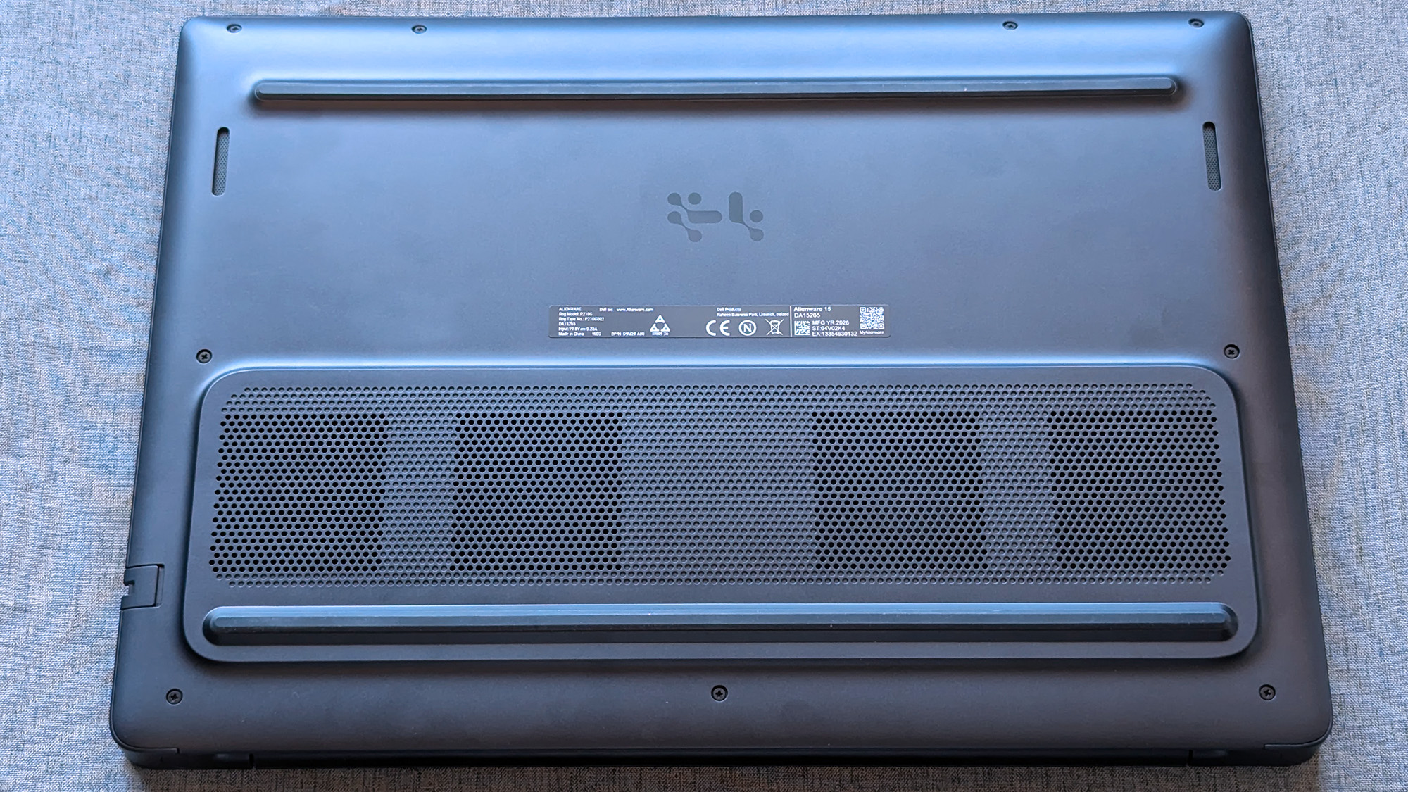 The underside of an Alienware laptop rests on a fabric surface, showing ventilation grilles, rubber feet, and a central label with certification markings and a QR code.