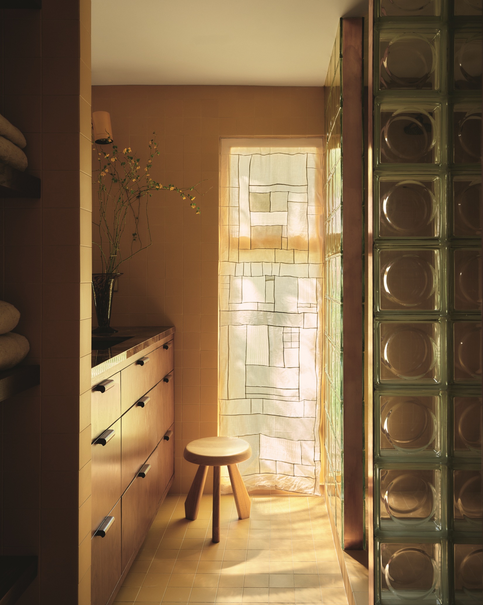 patchwork window covering in yellow kitchen with timber drawers, tiled floor, and glass brick wall with stool