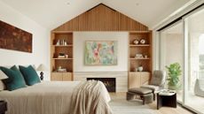 A bedroom with a vaulted ceiling, a wooden storage shelving unit, a sofa seater and footstool, a large bed draped in neutral bedlinen, a bobbin lamp by the bedside table and art overhead