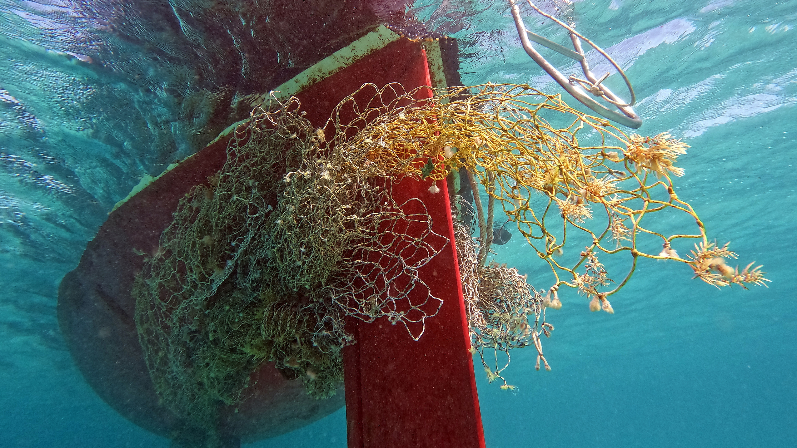 An underwater view of a red boat hull and keel entangled in a large, messy mass of discarded yellow and brown fishing nets in clear blue water.