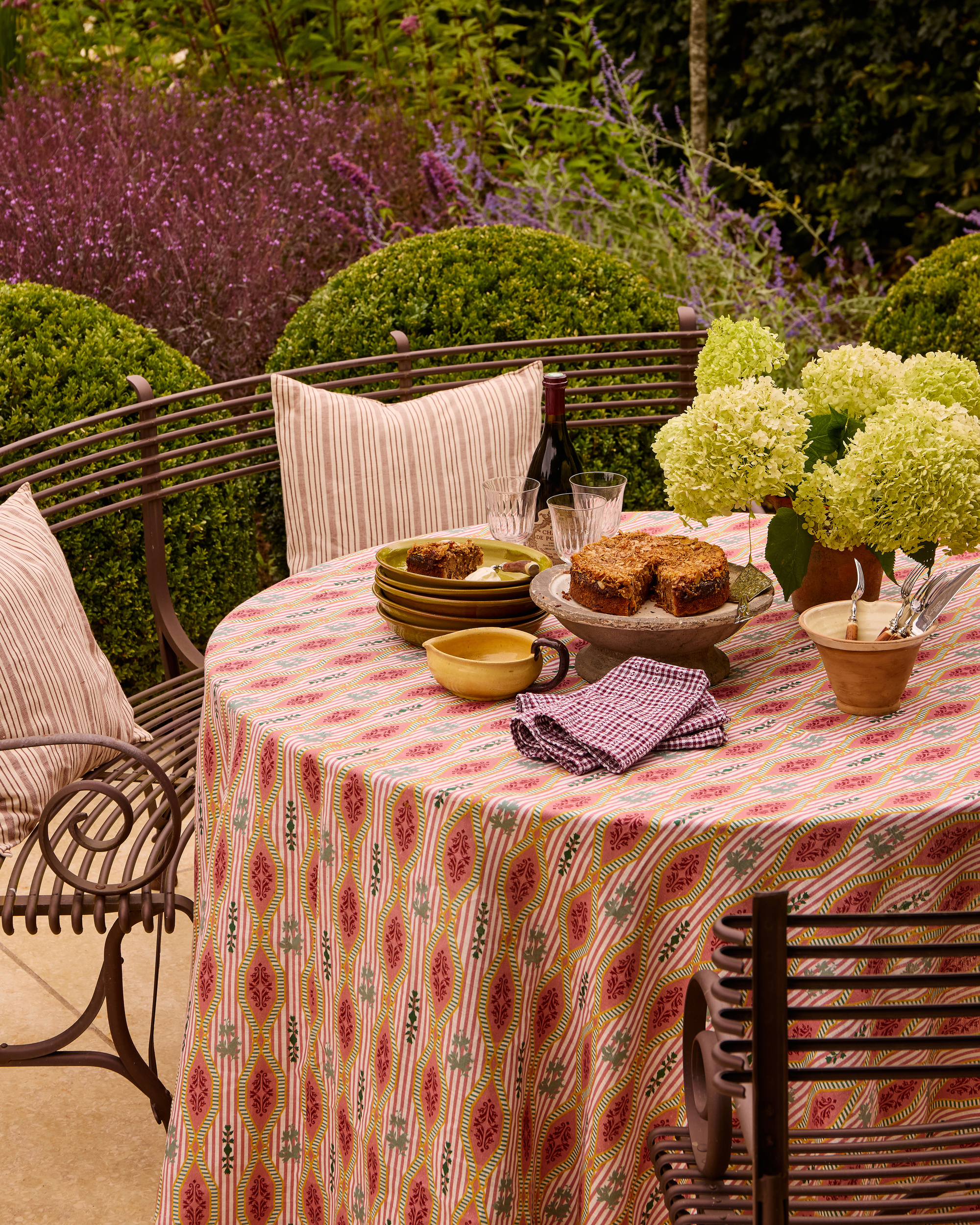 an outdoor table set with a patterned tablecloth, check napkins, a vase of hydrangeas, and cake on a platter