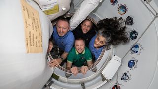 three men and a women pose inside the hatch of a white-walled space station