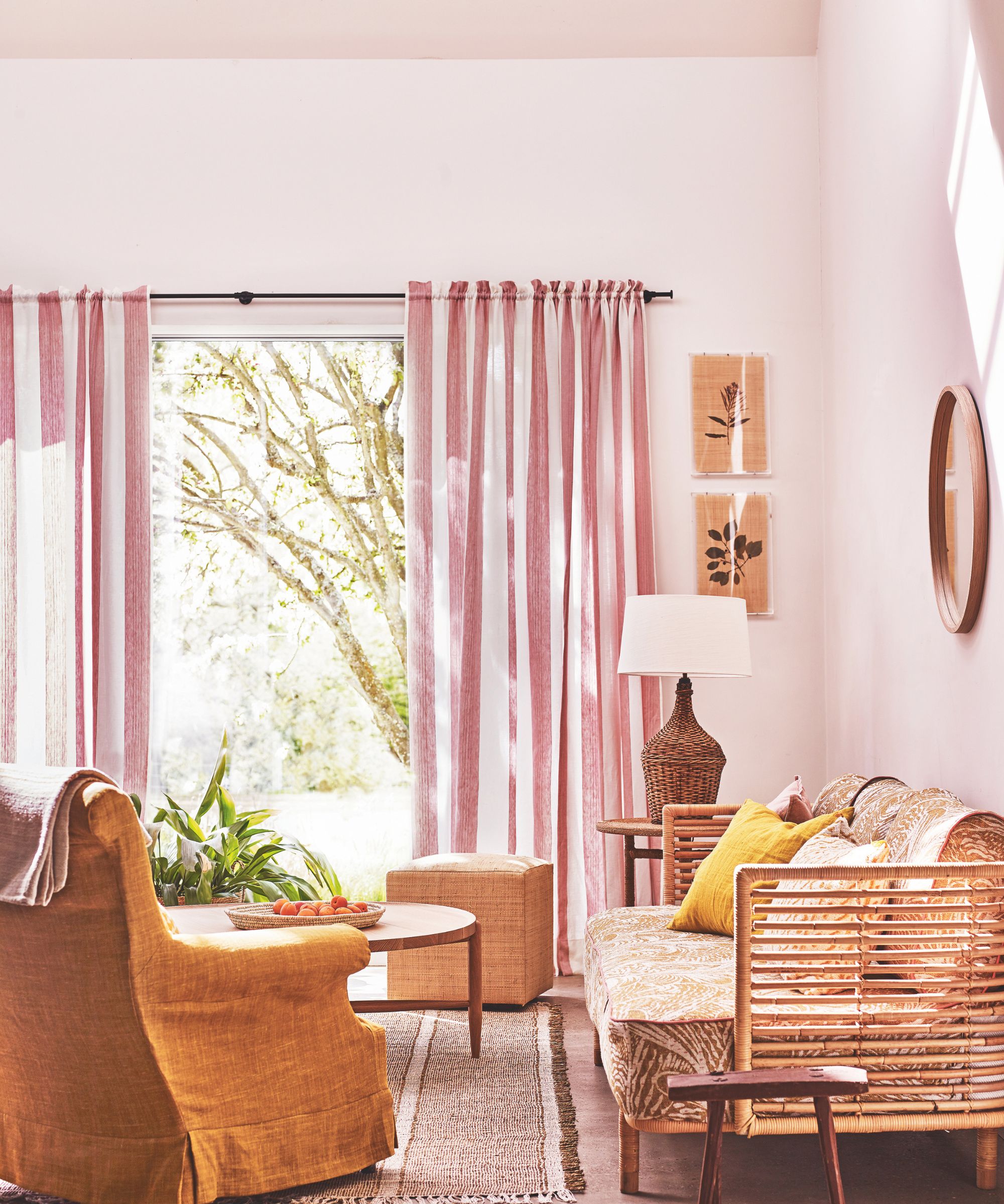 A pink and sunny color drenched living room with a wooden rattan sofa, white and pink striped curtains and a burnt ochre chair. On the floor is a brown and white striped rug and a circular wooden table.