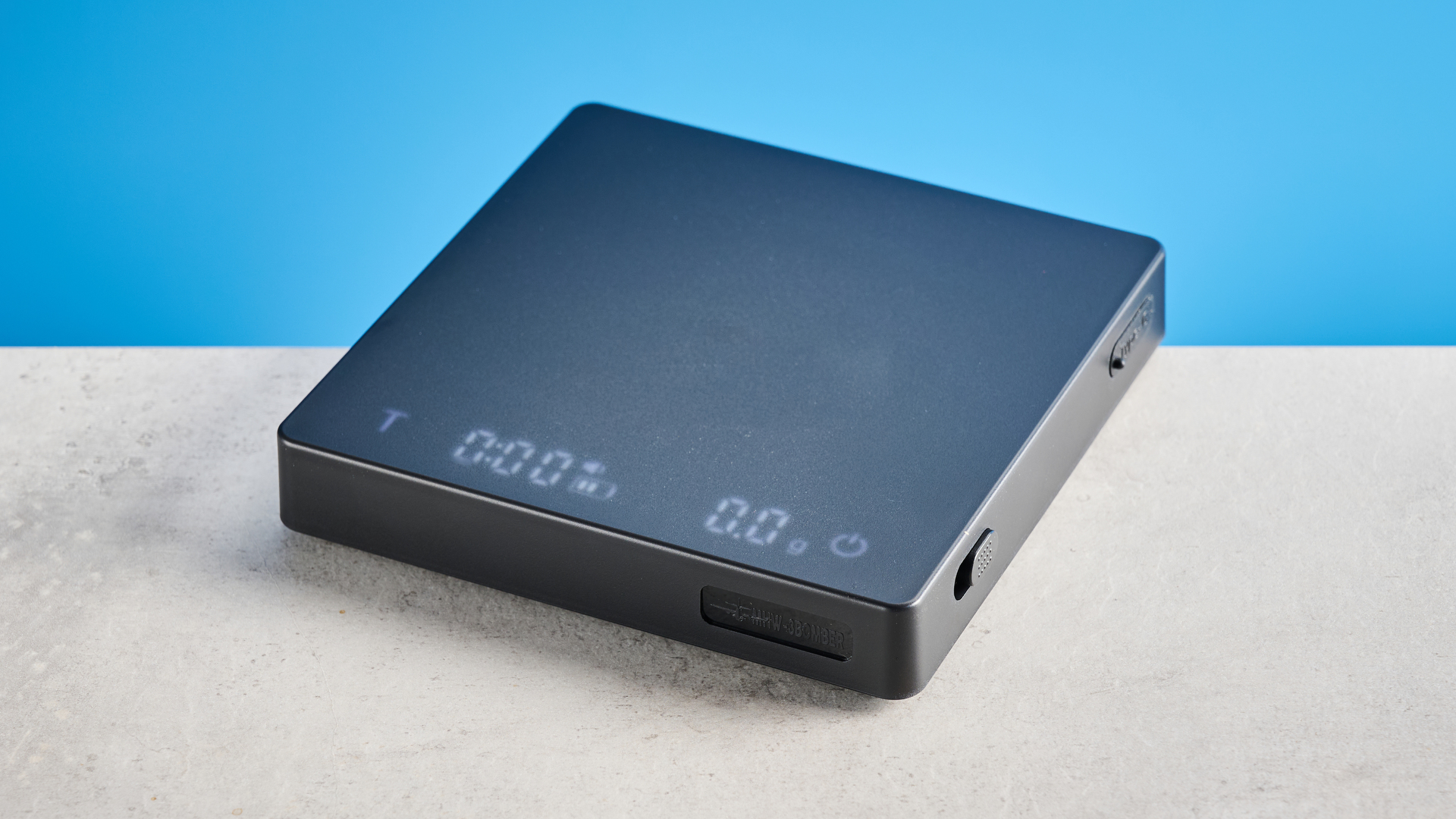 the mhw-3bomber cube coffee scale 2.0 in black photographed against a blue background showing its button controls and dial surface