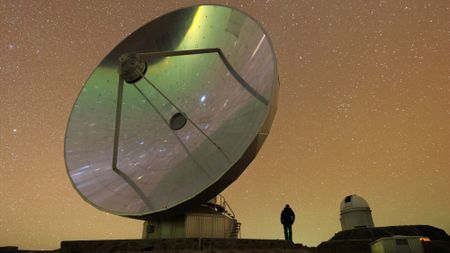 A large telescope dish points skywards against the backdrop of a starry night sky infused with an orange glow. A man stands in the foreground and stars can be seen reflected in the telescope dish.