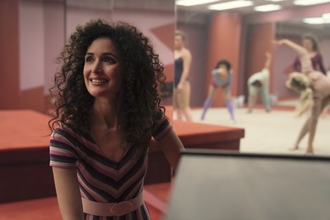 Sheila (Rose Byrne) in the aerobics studio, smiling, with permed hair and wearing a short-sleeved top with purple, green and pink chevrons. Next to her in the mirror we can see women in leotards warming up for an aerobics class.