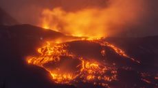 A volcano glowing with orange and yellow lava is seen against the evening night sky.
