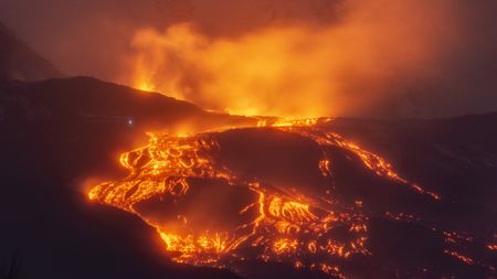 A volcano glowing with orange and yellow lava is seen against the evening night sky.