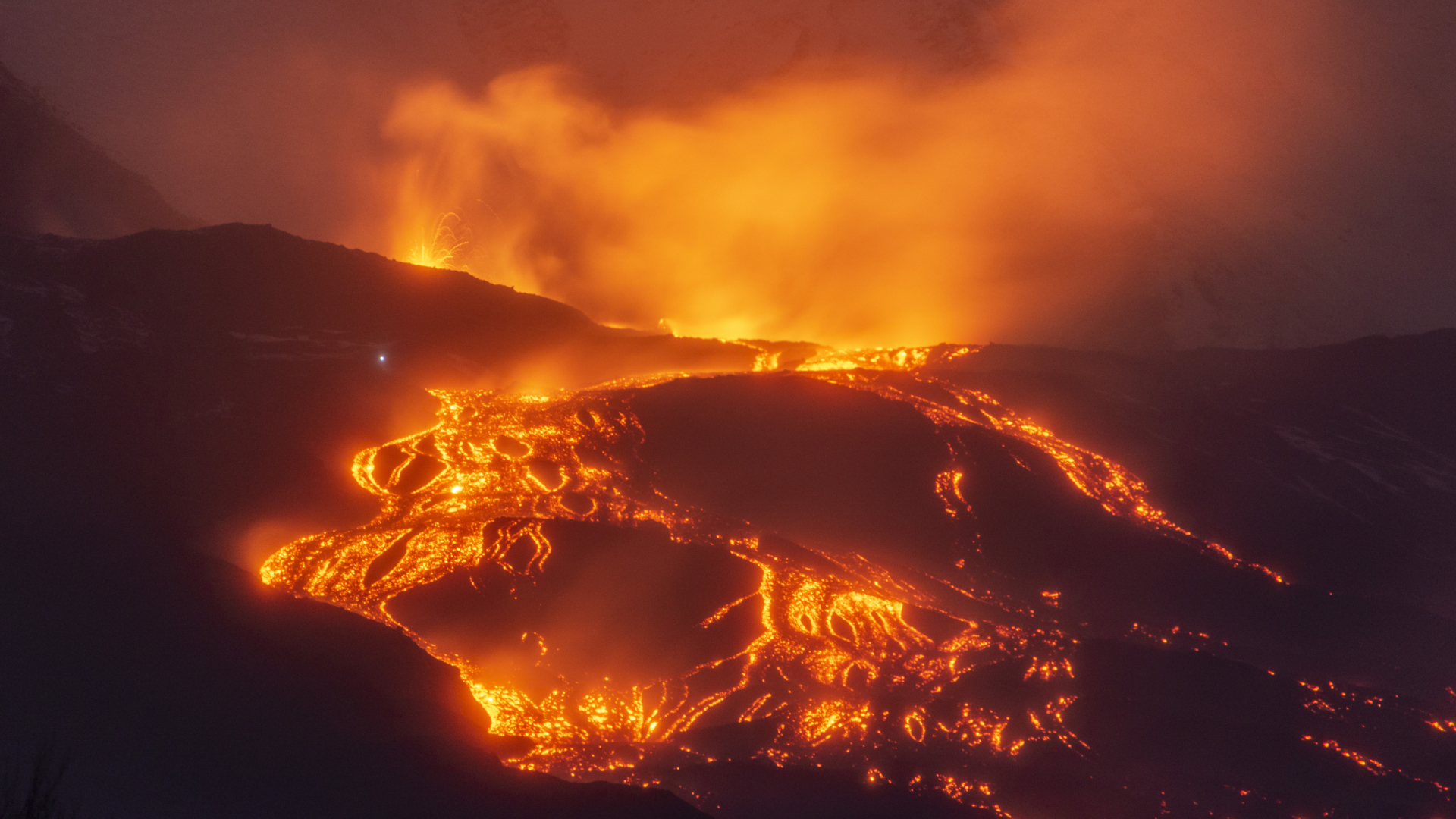 A volcano glowing with orange and yellow lava is seen against the evening night sky.