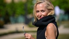 Gillian Anderson at the Jacquemus: Photocall - Paris Fashion Week Menswear Spring/Summer 2026 in Versailles, pictured outside in a sunny garden with a black high neck dress on, smiling side on to the camera with sunglasses in her hand