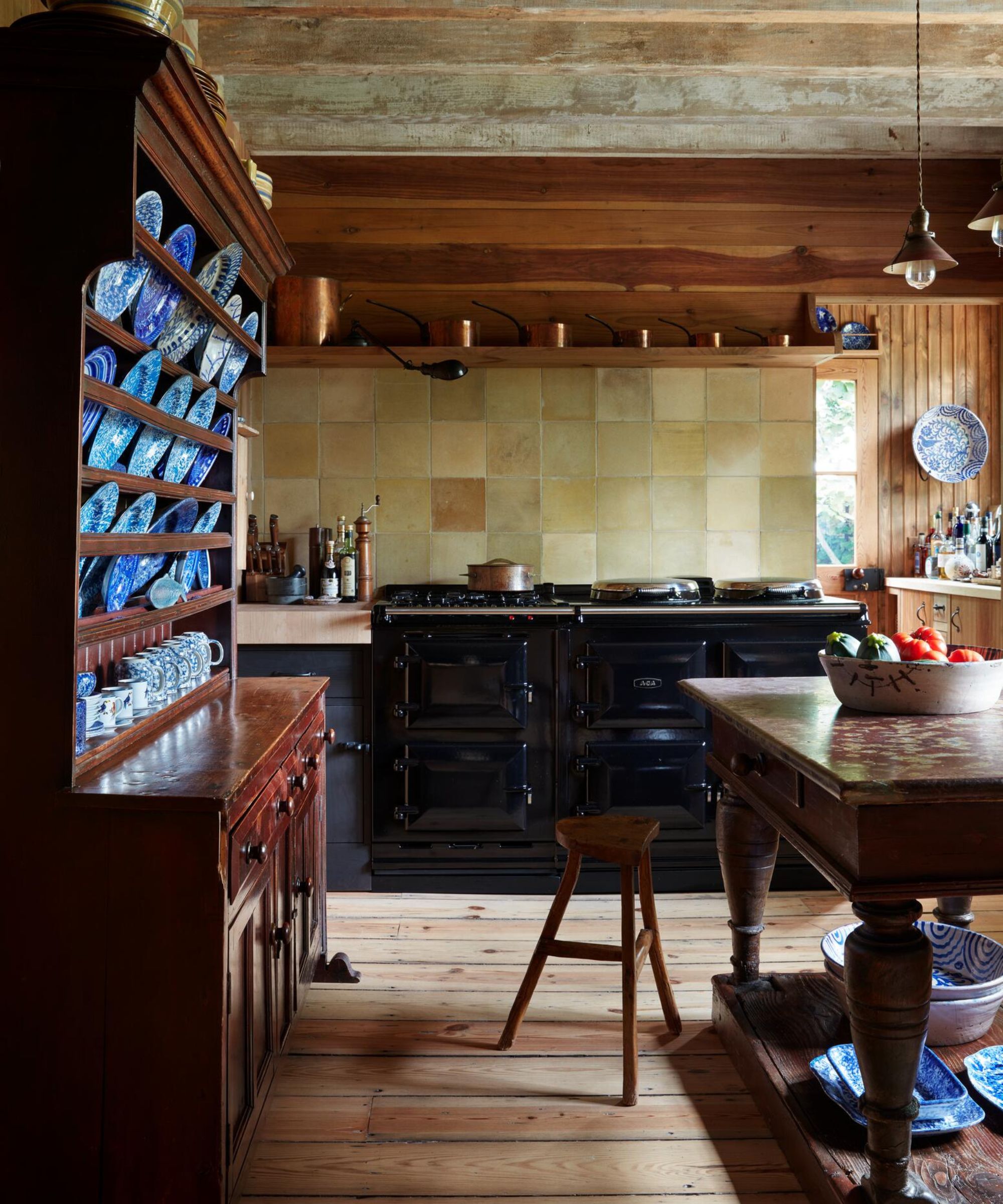 A rustic kitchen with rich wood walls and ceilings. It features a large black Aga-style range set against tan square tiles, a tall antique hutch filled with blue and white patterned plates, and a heavy wooden island in the foreground.