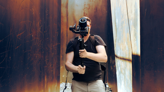 A photographer holding a camera on a gimbal camera in front of their body, surrounded by industrial metallic panels with scratched graffiti