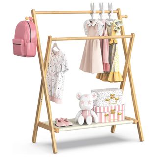 A wooden kids clothing rail with two racks and a shelf with dresses, pink backpack, and a shelf with two patterned suitcases, a soft toy and a pair of pink shoes