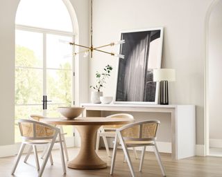 white dining room with large glass doors, round wooden table, rattan chairs and white console table with black artwork and lamp