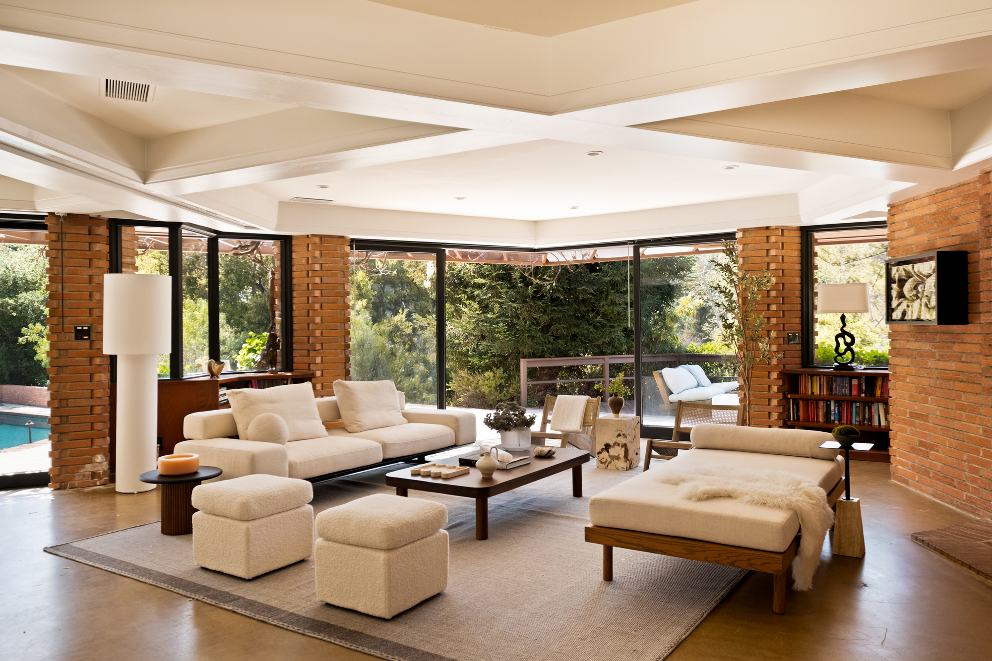 Image of an open concept living room that has windowed doors that open to the outdoors. There is a modern sofa, a modern chaise lounge, and two white ottomans around a wooden coffee table in the center of the room.