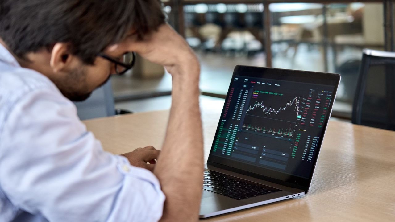 An investor looks frustrated as he looks at a trading graph on his laptop at a conference room table.