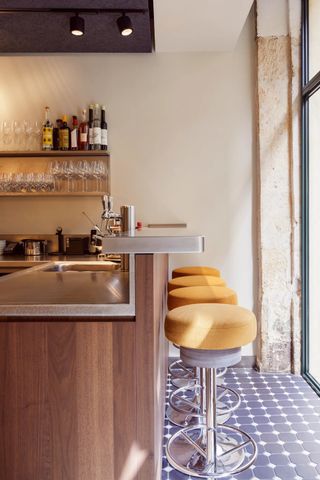 A modernly furnished restaurants with tile floors, chrome and yellow velvet tall stools, a wooden and iron bar counter, and industrial lighting is captured in natural light.