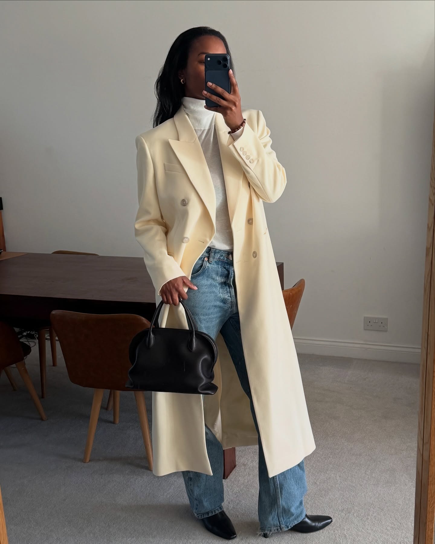 @nlmarilyn wearing a long cream coat, white turtleneck, and light-wash jeans with black boots and a black The Row bag.