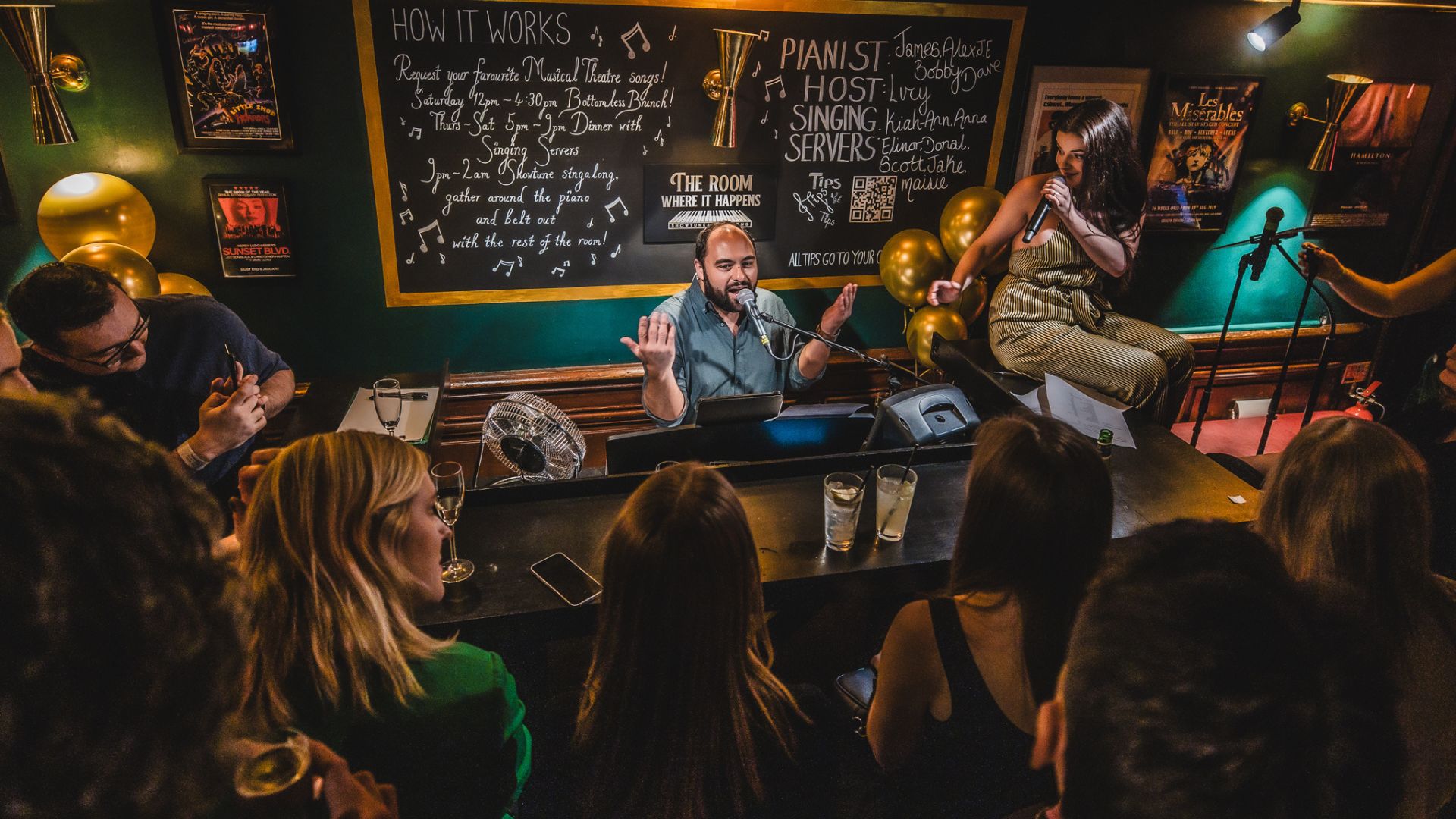 an image of the pianist singing whilst one of the servers crouches on the piano singing a duet with him whilst a crowd surrounds them