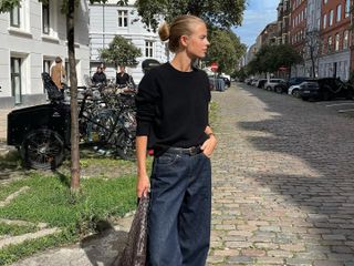 Art director and fashion influencer Clara Dyrhauge on the street in Europe wearing dark jeans, a black belt, a black short-sleeve knit top, black pointed-toe Aeyde shoes, and carrying a woven handbag.