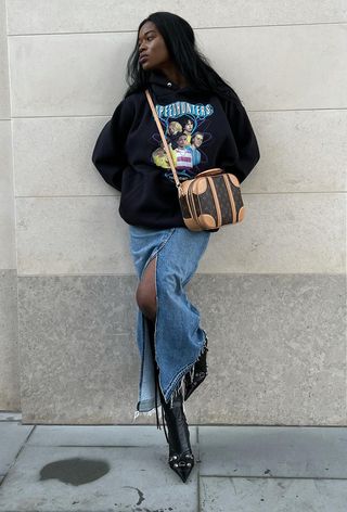 A woman standing outside wearing a black graphic hoodie with a denim maxi skirt, black knee-high boots, and a crossbody bag.